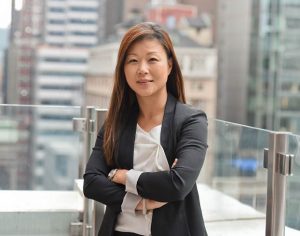 Mimi Song standing on a rooftop with arms crossed, wearing a black blazer, with city buildings in the background.