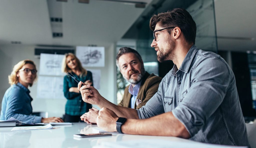 Team of four professionals in a meeting room, with one man speaking while others listen and take notes during a discussion.