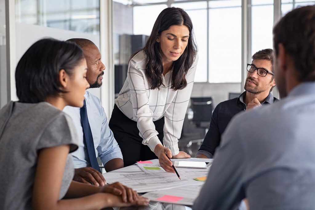 Women leading a team discussion around a table with documents and sticky notes, as four colleagues listen attentively.