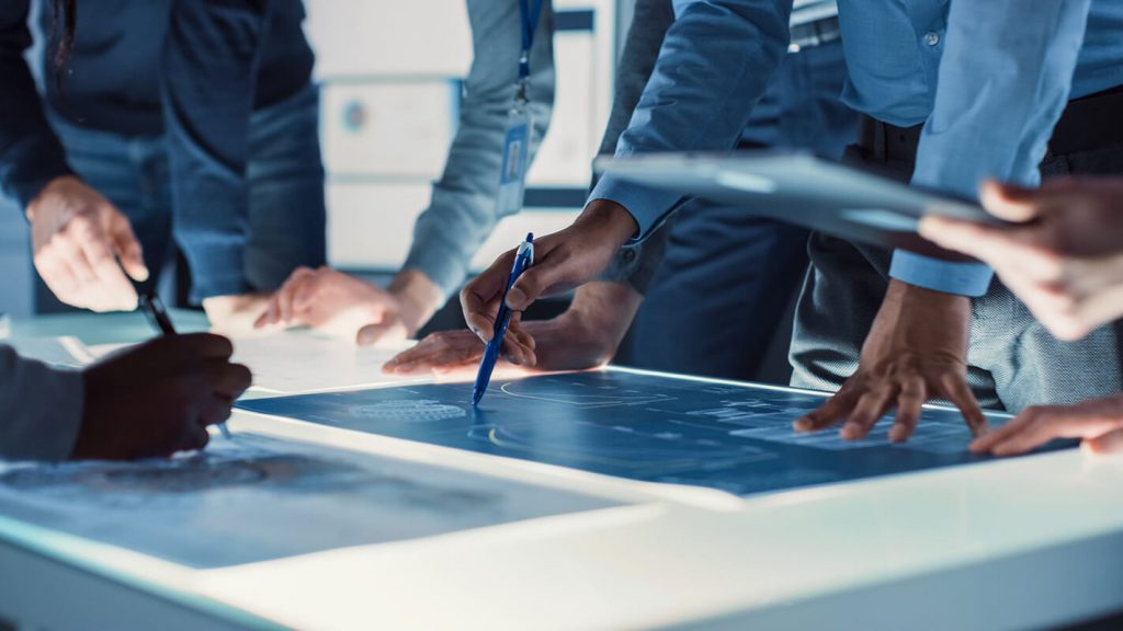 People collaborating over blueprints on a lit table, with several hands pointing and writing, wearing business attire.