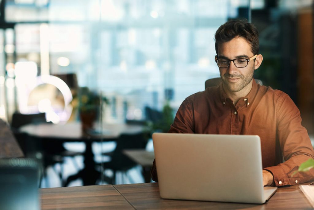 Man in a brown shirt working on a laptop at a wooden desk in a modern office with large windows.