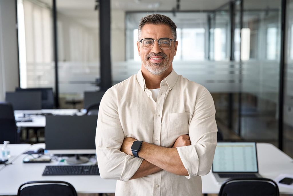 Confident man wearing glasses and a smart casual shirt standing with arms crossed in a modern office with computer desks behind him.