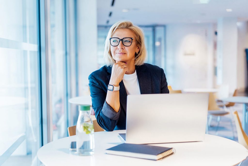 A person with blond hair and glasses sitting at a round table in a bright office, with a laptop, notebook, and water bottle in front.