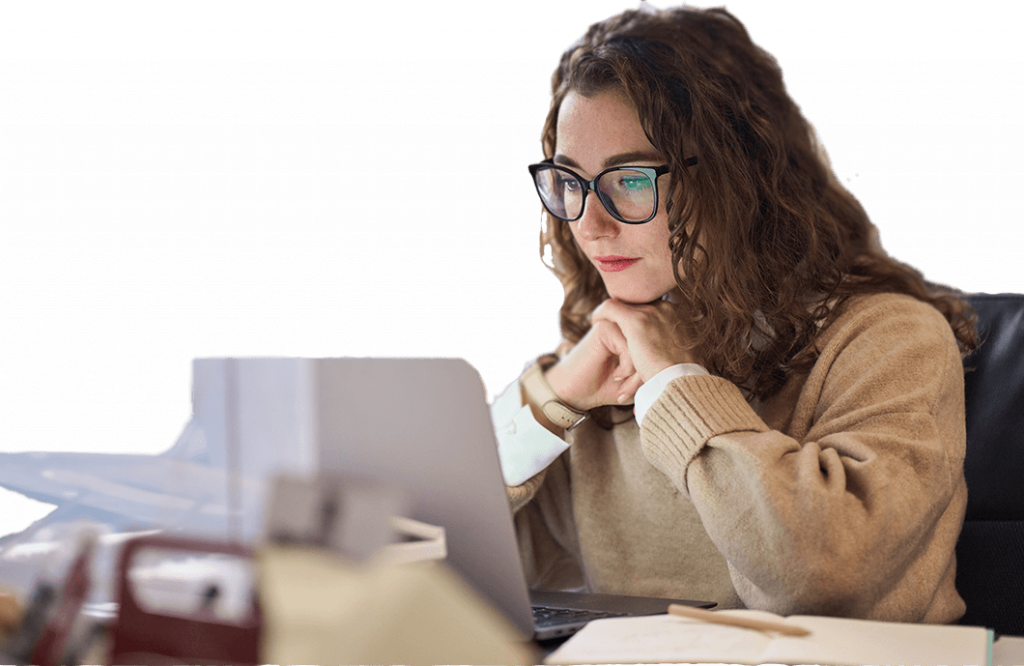 Woman with curly hair and glasses focused on a laptop, with papers nearby, sitting in an office environment.