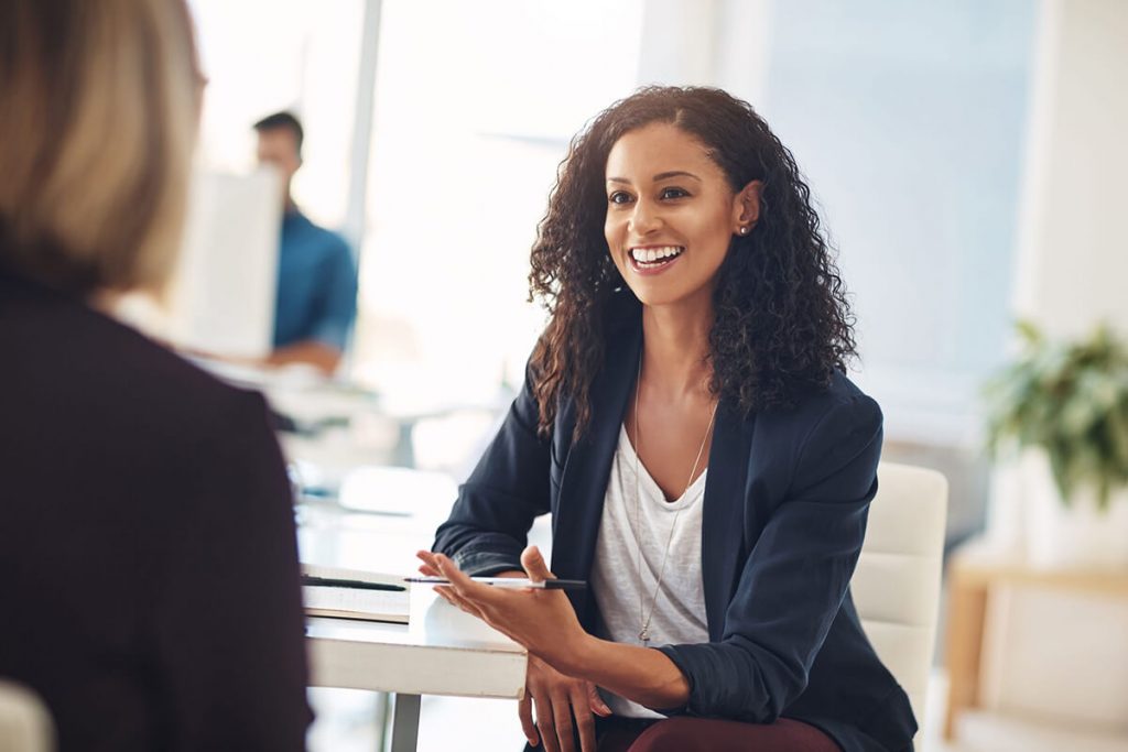 A woman with curly hair smiles while talking to someone across a desk in an office setting.
