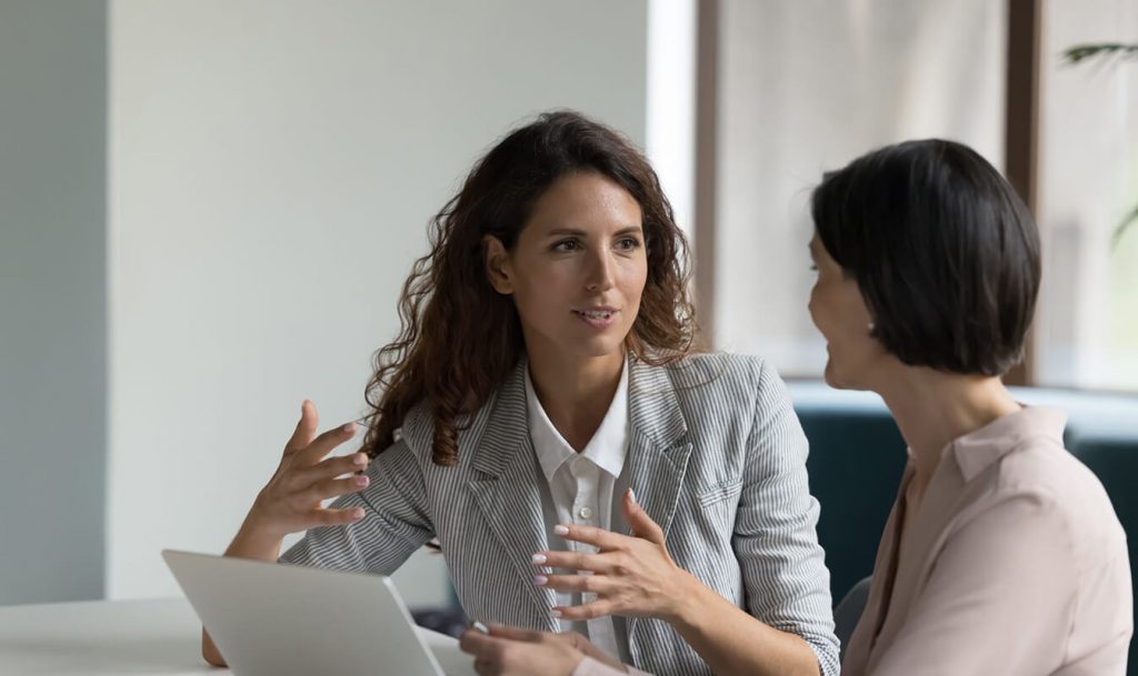 Two women seated at a desk, engaging in a discussion, with a laptop in front of them. Bright room with large windows.