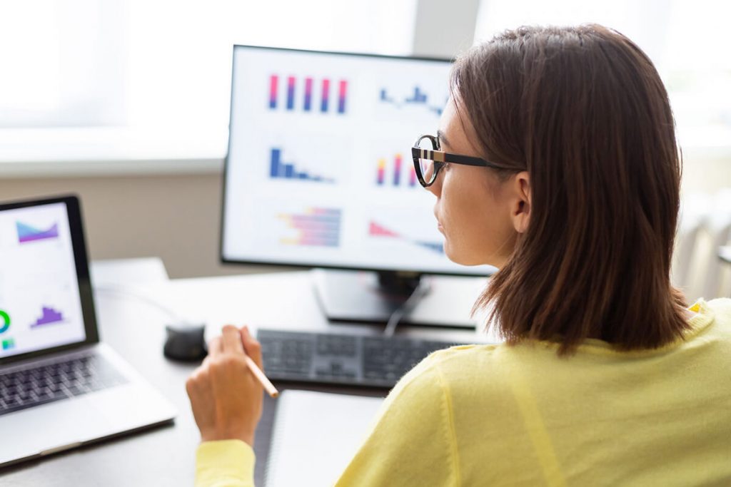 A person in glasses is analyzing graphs on computer screens, with a laptop beside them on a desk.