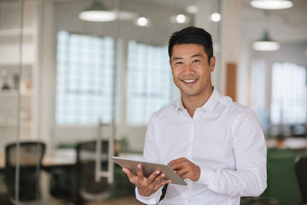 Smiling man in white shirt holding a tablet in a bright office with glass walls and modern furniture.