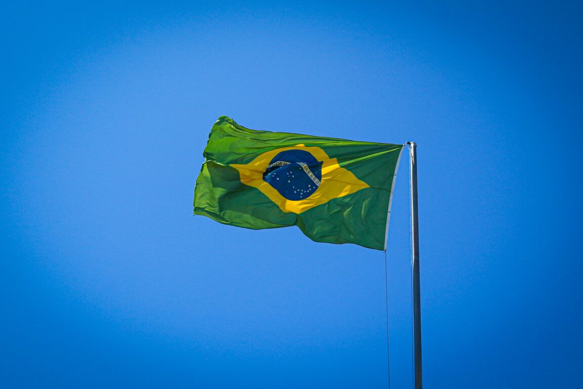 The Brazilian flag waves in a bright blue sky. The flag features a green field wiht a large yellow diamond in the center. Inside the diamond is a blue globe with 27 white stars arranged to depict the night sky over Rio de Janeiro, as it appeared on November 15, 1989, the date of the proclamation of the Republic.
